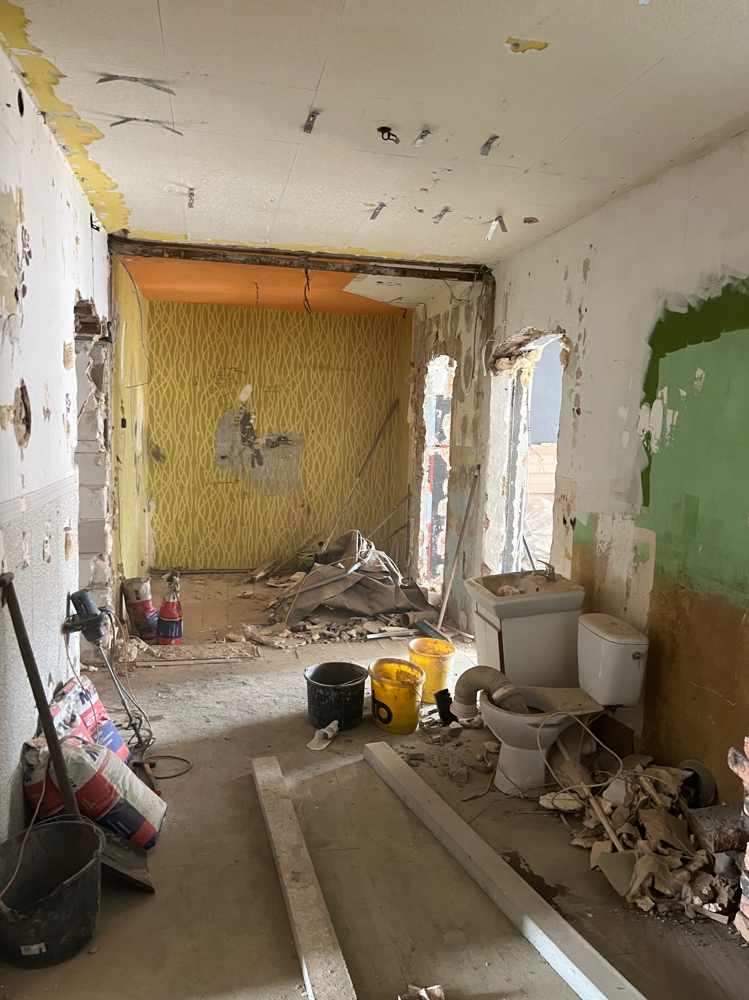 Wnętrze mieszkania w trakcie generalnego remontu, widoczne ślady po zerwanych tapetach, resztki gruzu, odsłonięte instalacje sufitowe, częściowo zdemontowana toaleta i umywalka, worki z zaprawą...