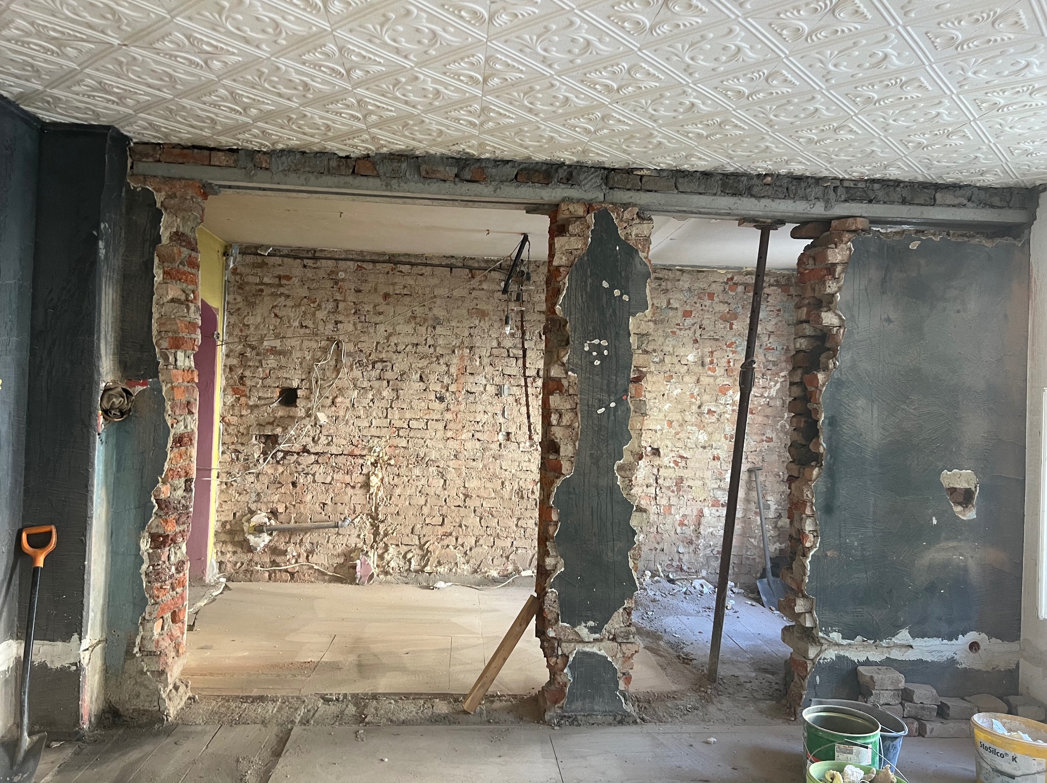 Zdjęcie przedstawia wnętrze w trakcie demontażu ścian działowych z odsłoniętą cegłą, widoczne podparte stalowe belki stropowe, fragment dekoracyjnego sufitu kasetonowego, oraz narzędzia pracy...