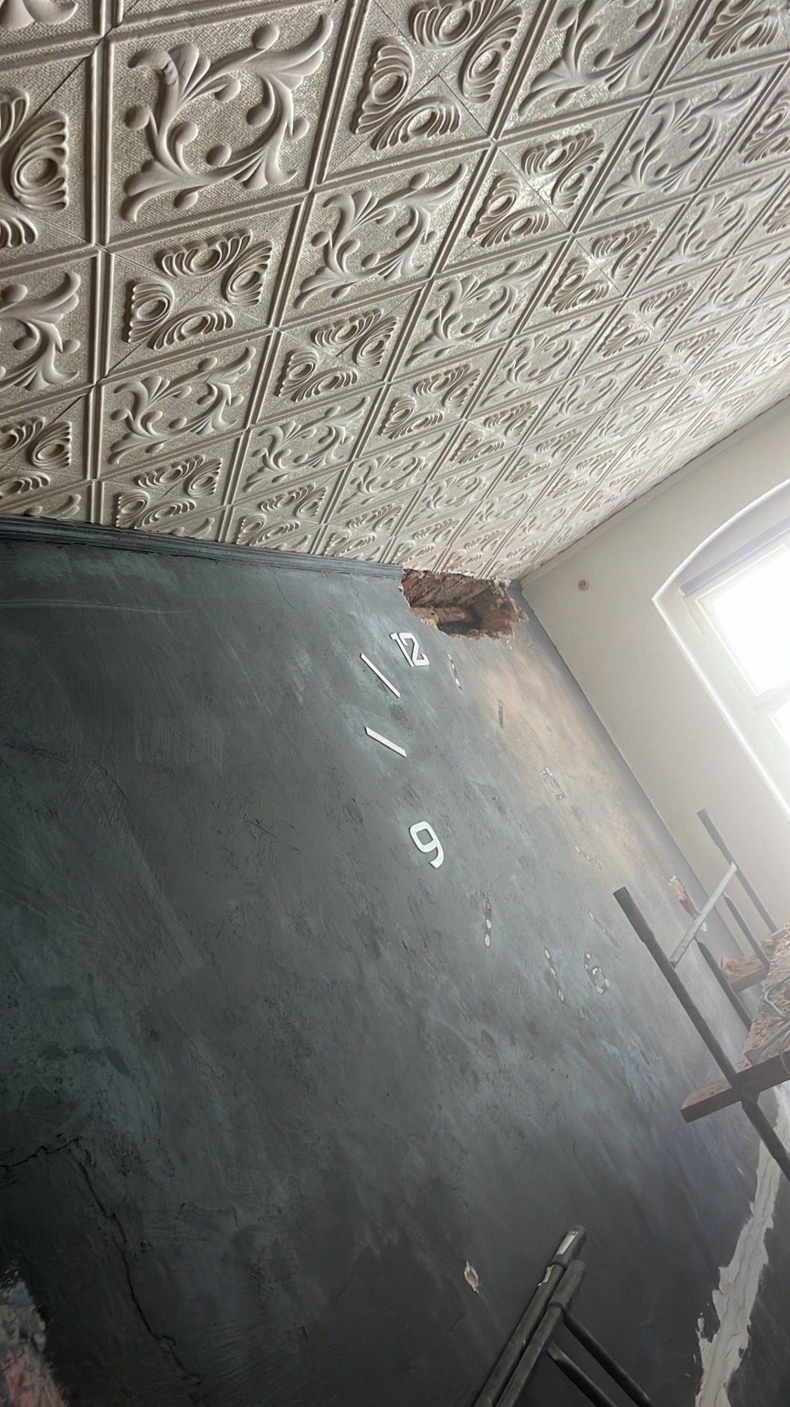 Ściana z efektem dekoracyjnym imitującym beton, z naklejonymi cyframi '12' i '9' tworzącymi zegar, widoczny ubytek tynku przy suficie i ozdobny sufit kasetonowy.