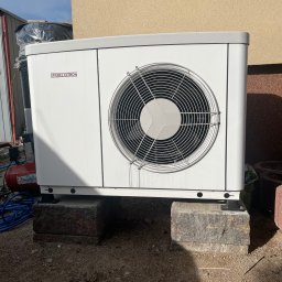 NEWBUD PRO - Biała pompa ciepła marki Stiebel Eltron, zamontowana na betonowych blokach przy ścianie budynku. Widoczne przewody i elementy instalacji.