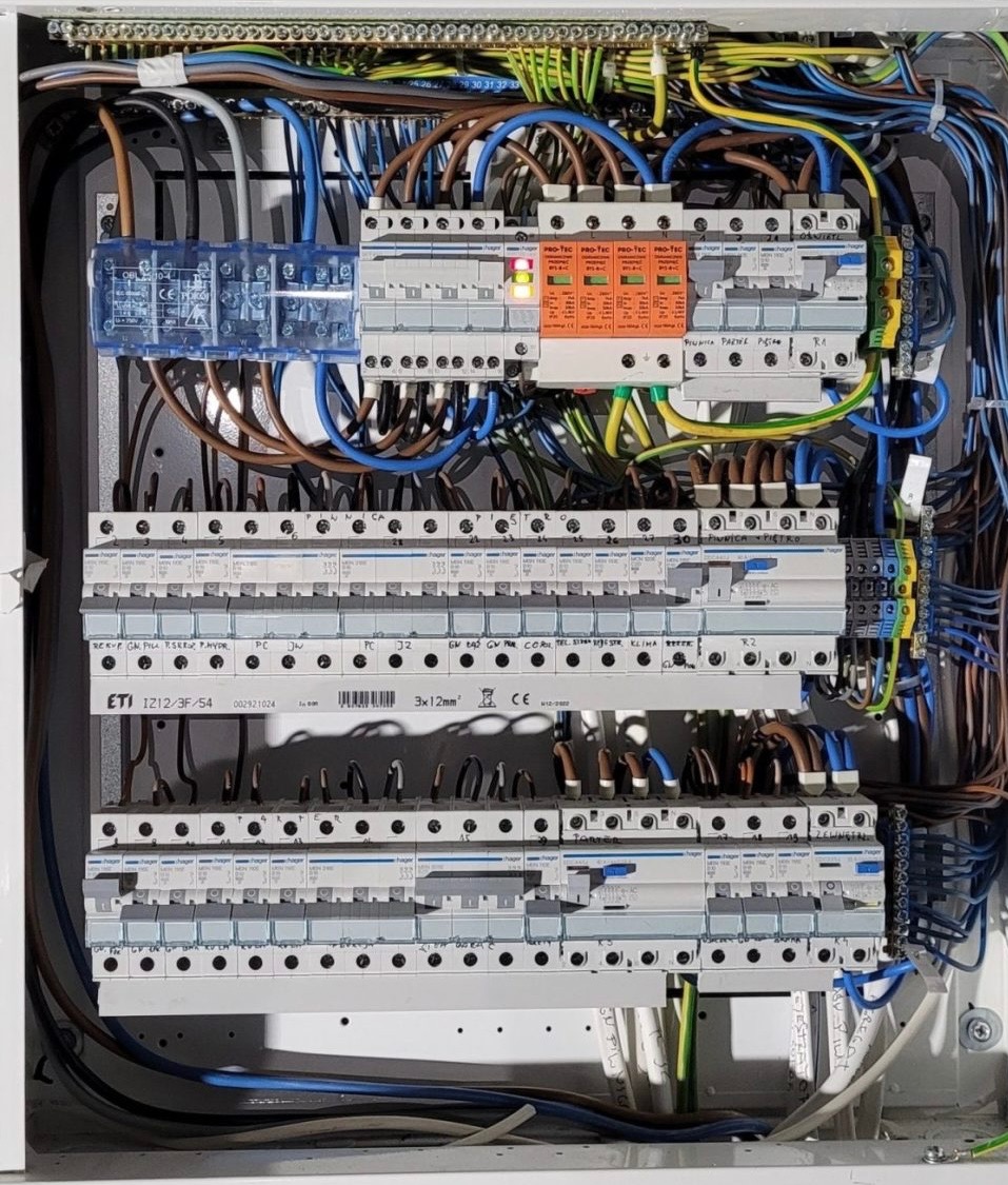 Rozdzielnia elektryczna z widocznymi zabezpieczeniami, przewodami i aparaturą modułową. Detal instalacji z pomarańczowym ogranicznikiem przepięć i niebieskim modułem.