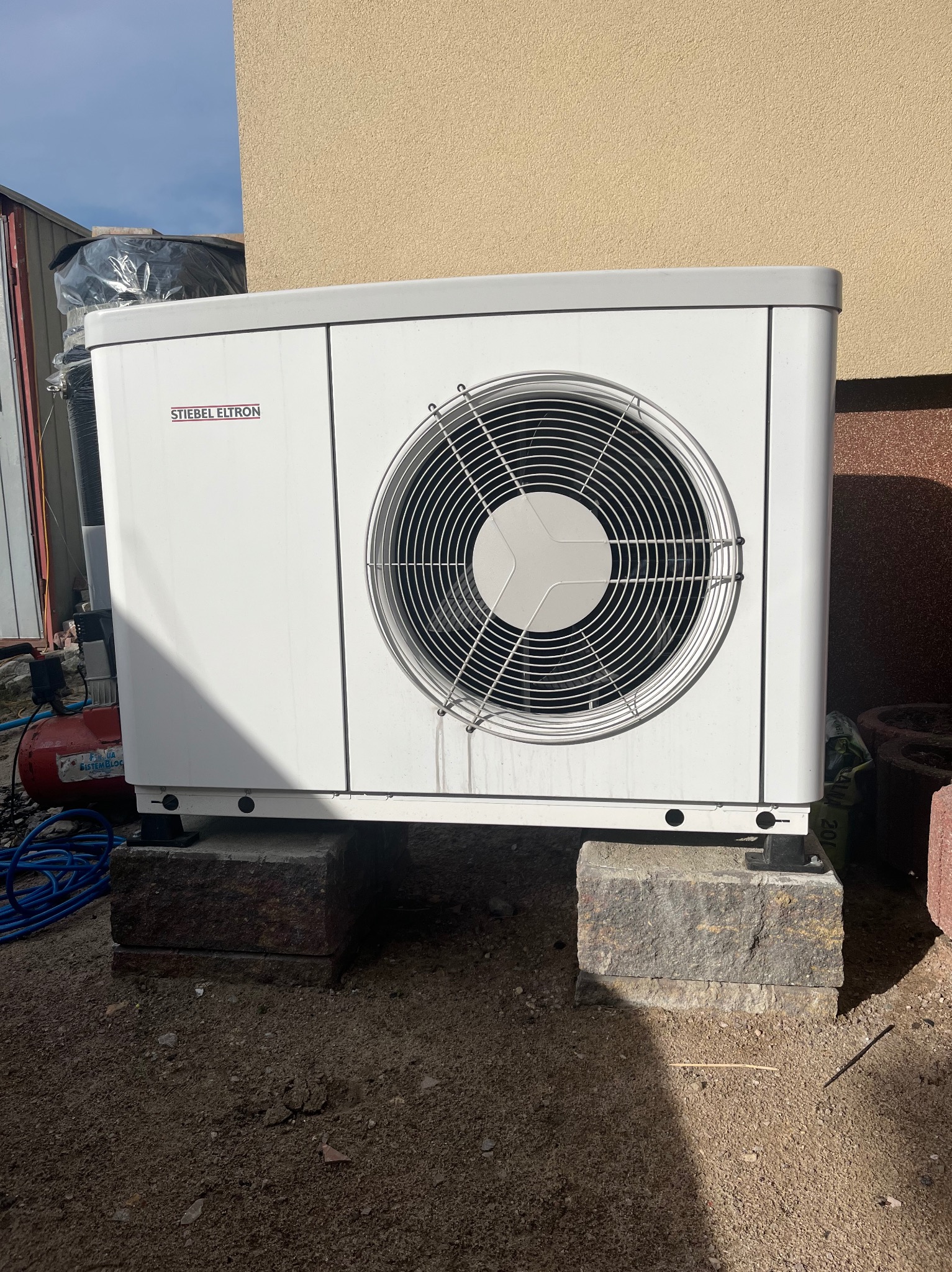 Biała pompa ciepła marki Stiebel Eltron, zamontowana na betonowych blokach przy ścianie budynku. Widoczne przewody i elementy instalacji.