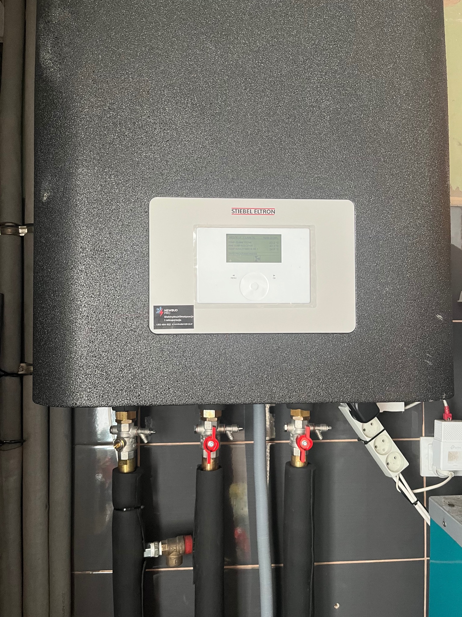 Szary piec Stiebel Eltron z widocznym panelem sterowania i instalacją hydrauliczną. Na ścianie czarne płytki, obok gniazdko elektryczne.