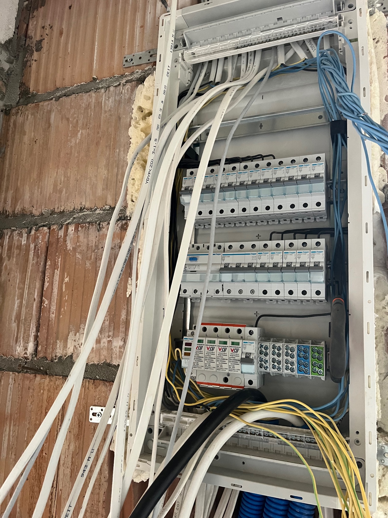 Rozdzielnica elektryczna z licznymi przewodami w trakcie montażu na ścianie z cegły. Widoczne zabezpieczenia i listwy zaciskowe. Prace instalacyjne w surowym wnętrzu.