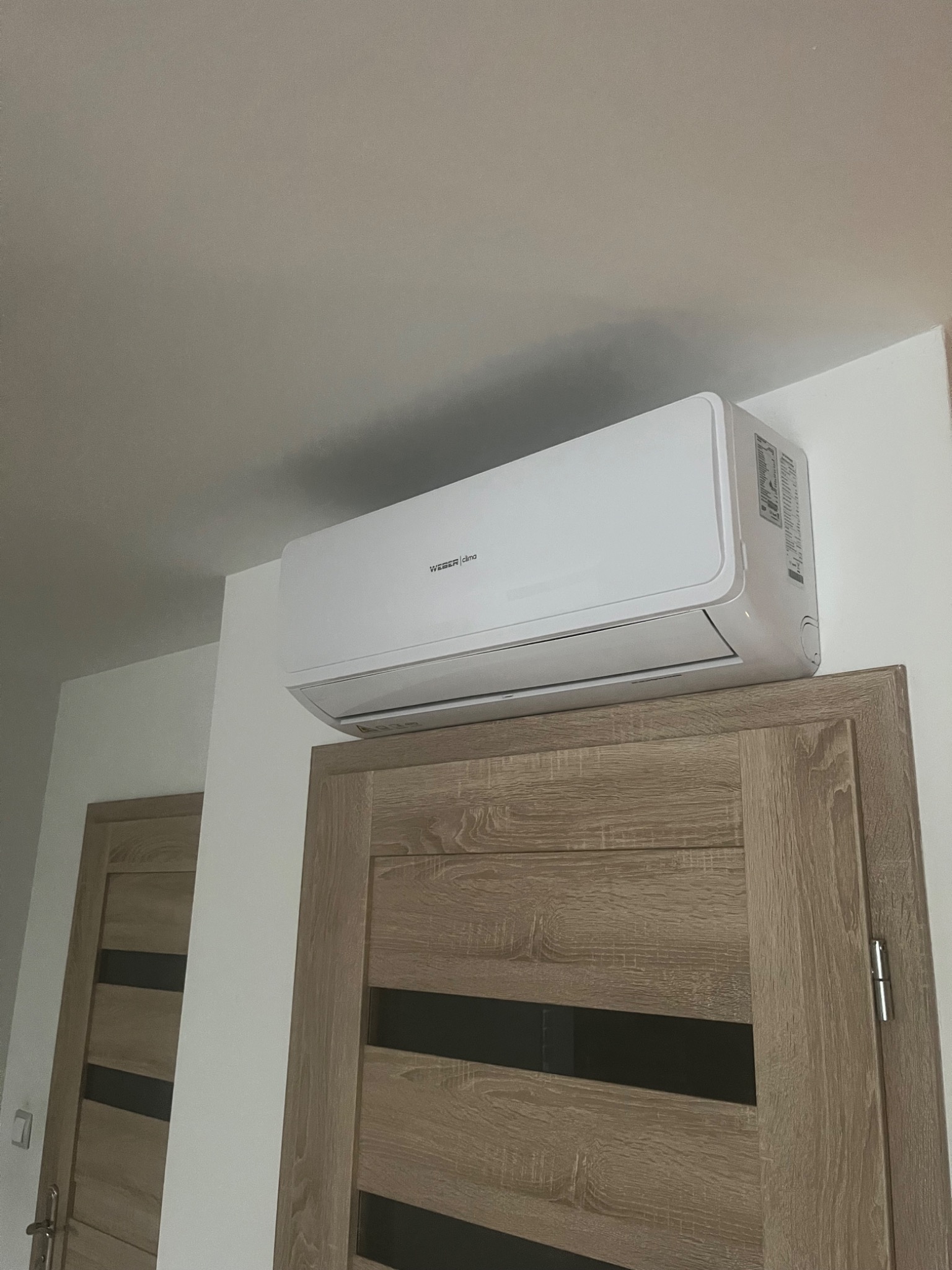 Biała klimatyzacja WEBER clima zamontowana nad drewnianymi drzwiami z czarnymi wstawkami, widok od dołu.