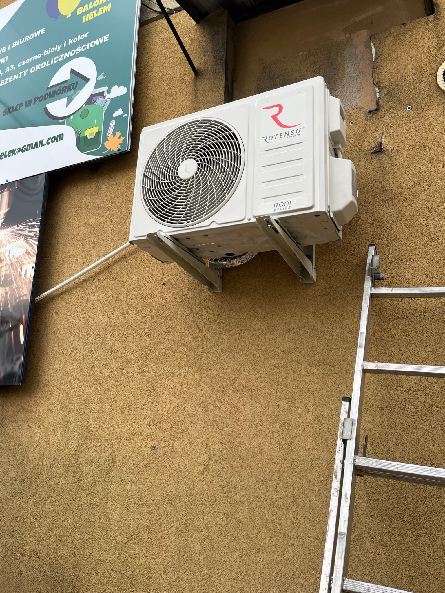 Zamontowana klimatyzacja Rotenso Roni na elewacji budynku, widoczna drabina. Instalacja z rurką odprowadzającą. Nad klimatyzacją szyld reklamowy.