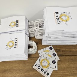 KOALA DRUK - Stosy białych toreb papierowych z nadrukiem logo Akademii Ruchu i Tańca POWER oraz naklejki z logo drukarni leżące na drewnianym stoliku.