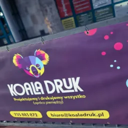 Fioletowy baner reklamowy z kolorową grafiką koali i hasłem 'KOALA DRUK Projektujemy i drukujemy wszystko (oprócz pieniędzy)' zawieszony na metalowym ogrodzeniu.
