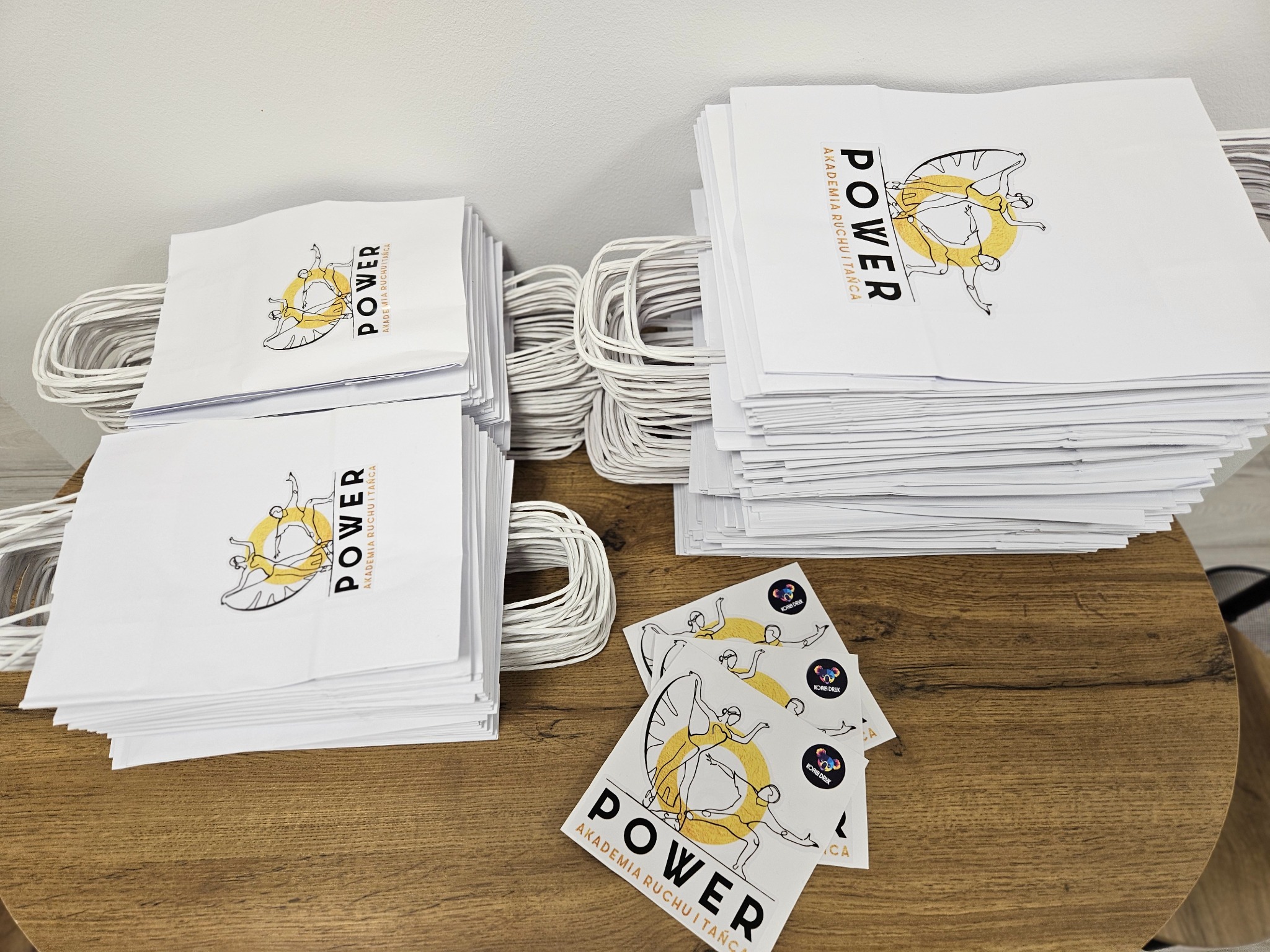 Stosy białych toreb papierowych z nadrukiem logo Akademii Ruchu i Tańca POWER oraz naklejki z logo drukarni leżące na drewnianym stoliku.