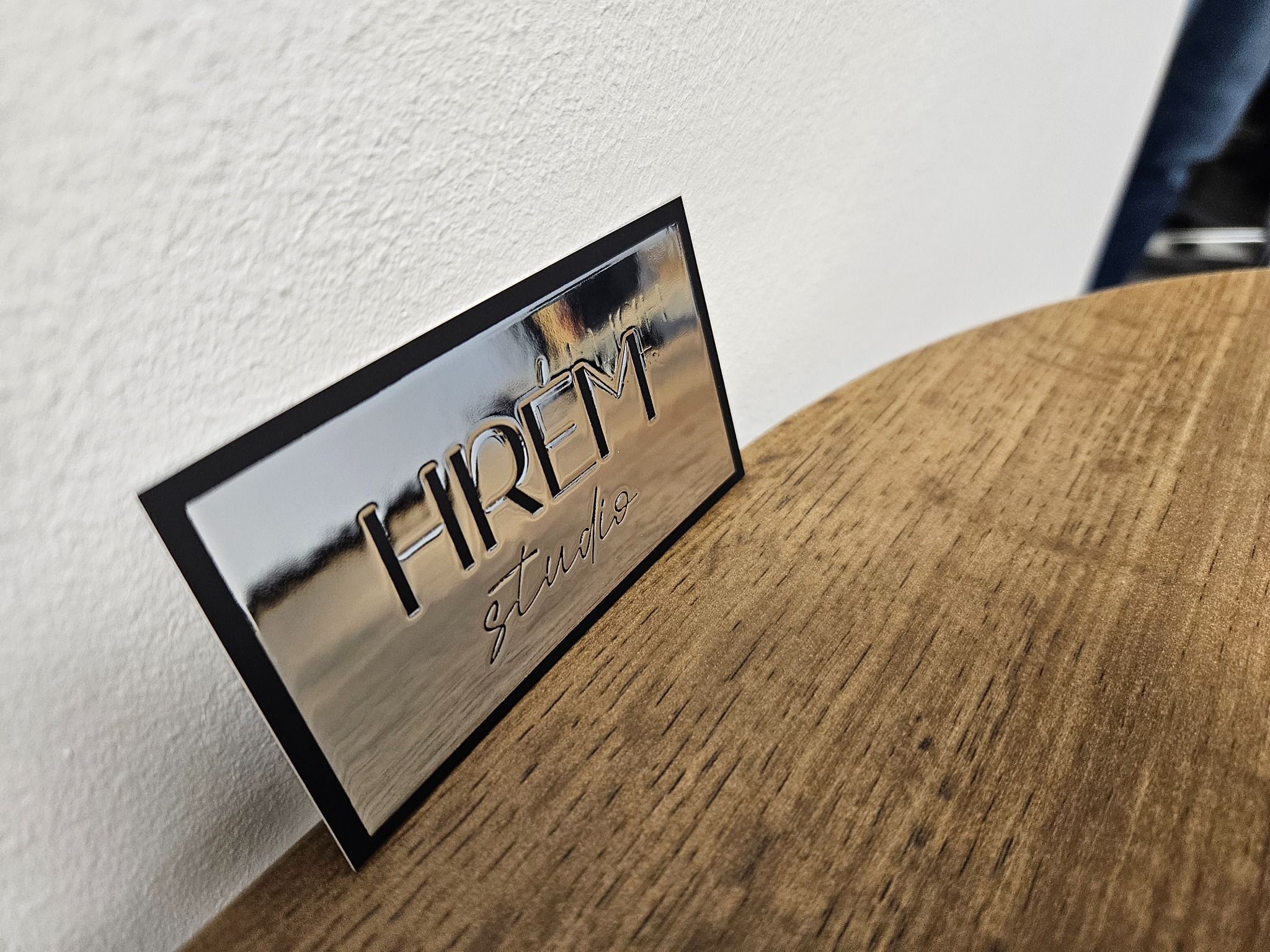 Błyszcząca wizytówka z czarną obwódką i wytłoczonym napisem 'Hirem studio' leży na drewnianym blacie, odbijając światło.