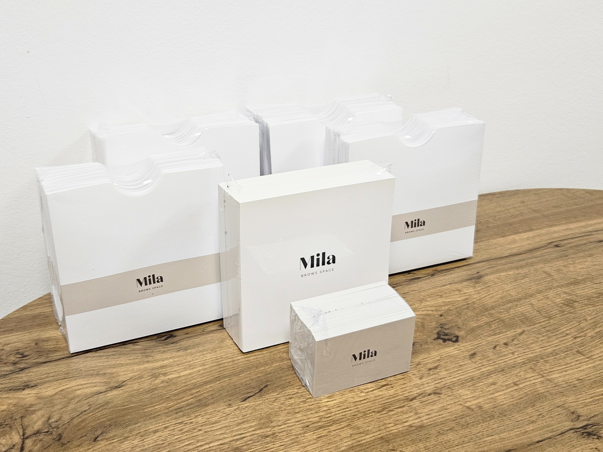 Stosy białych kartoników z logo 'Mila BROWS SPACE' na drewnianym stole, część w folii ochronnej.