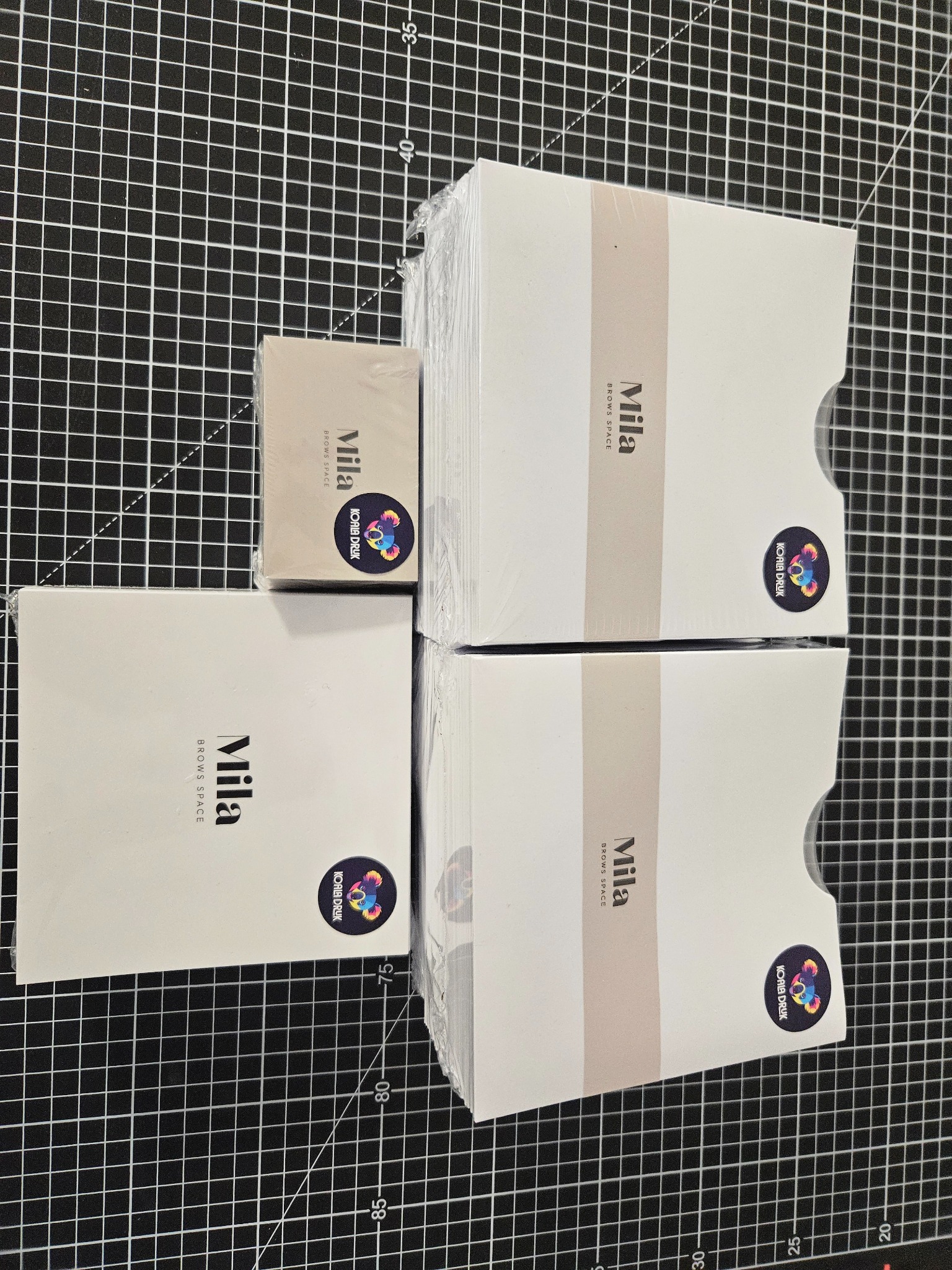 Stosy zapakowanych wizytówek z logo 'Mila Brows Space' i naklejką 'Koala Druk' na czarnym tle siatki pomiarowej.