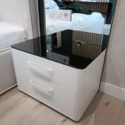 Biała szafka nocna z dwoma szufladami i czarnym, szklanym blatem, odbijająca fragment sypialni z łóżkiem i drewnianą ścianą.