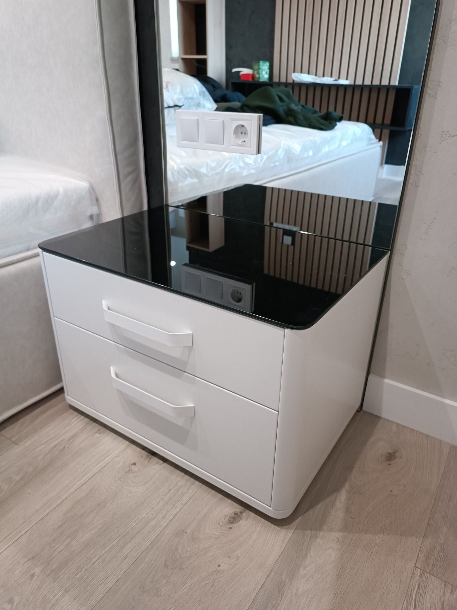 Biała szafka nocna z dwoma szufladami i czarnym, szklanym blatem, odbijająca fragment sypialni z łóżkiem i drewnianą ścianą.