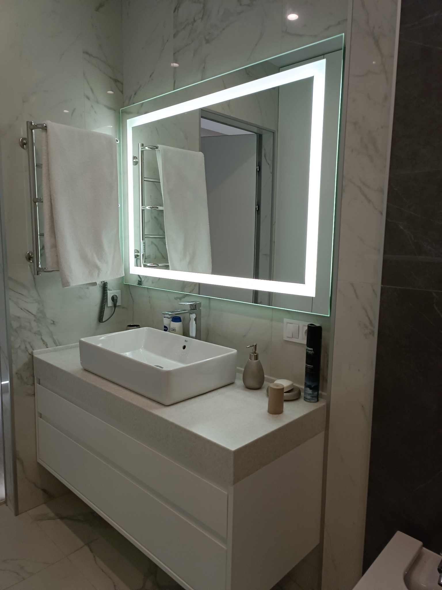 Nowoczesna łazienka z marmurowymi ścianami, podświetlanym lustrem, białą umywalką nablatową i szafką z szufladami.