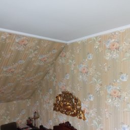 Wojciech Woroniecki - Pokój na poddaszu z tapetą w kwiaty, ukośny sufit i ozdobny, złoty ornament na ścianie.