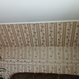 Wojciech Woroniecki - Beżowe tapety w pionowe pasy z motywem kwiatowym zdobią ściany i skośny sufit poddasza, widoczny fragment sofy i lampy.