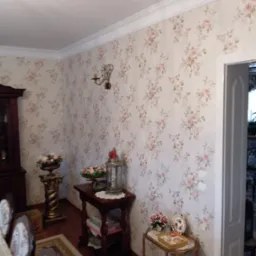 Pokój z tapetą w drobne róże, ozdobnymi listwami przysufitowymi, antykami i dekoracjami, widok na fragment jadalni z krzesłami w stylu retro