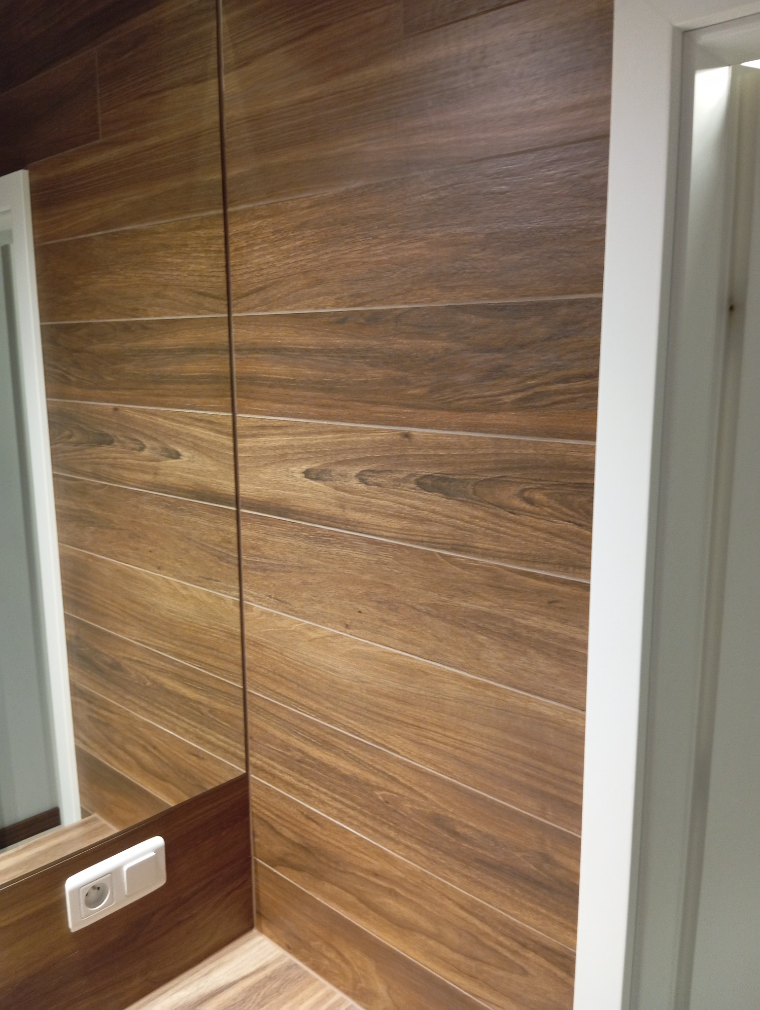 Ściana i podłoga wyłożone brązowymi płytkami imitującymi drewno w łazience, widoczne gniazdko elektryczne i biała ościeżnica drzwi.