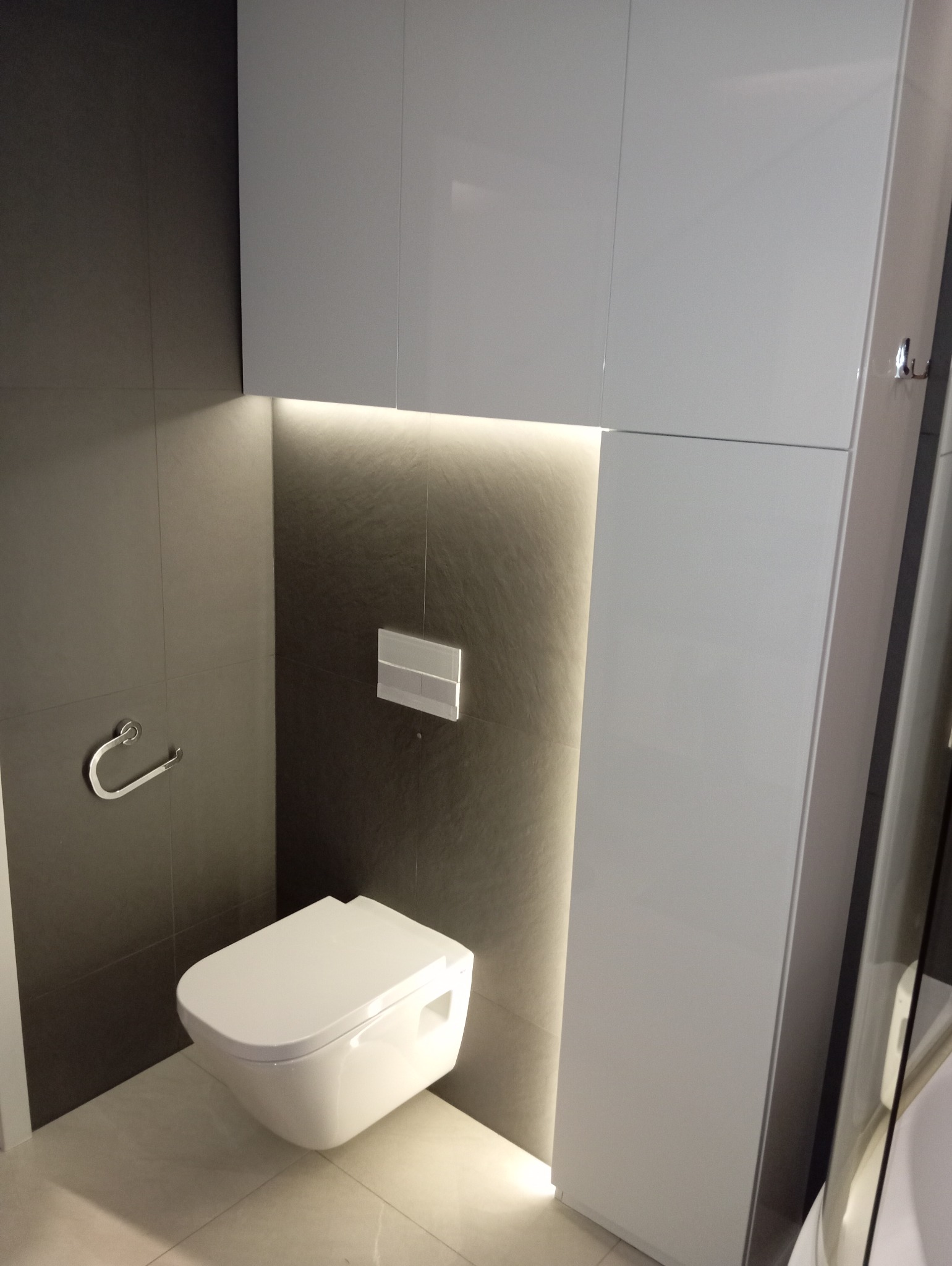 Nowoczesna toaleta podwieszana w łazience z szarymi płytkami i białymi szafkami, oświetlenie LED za toaletą.