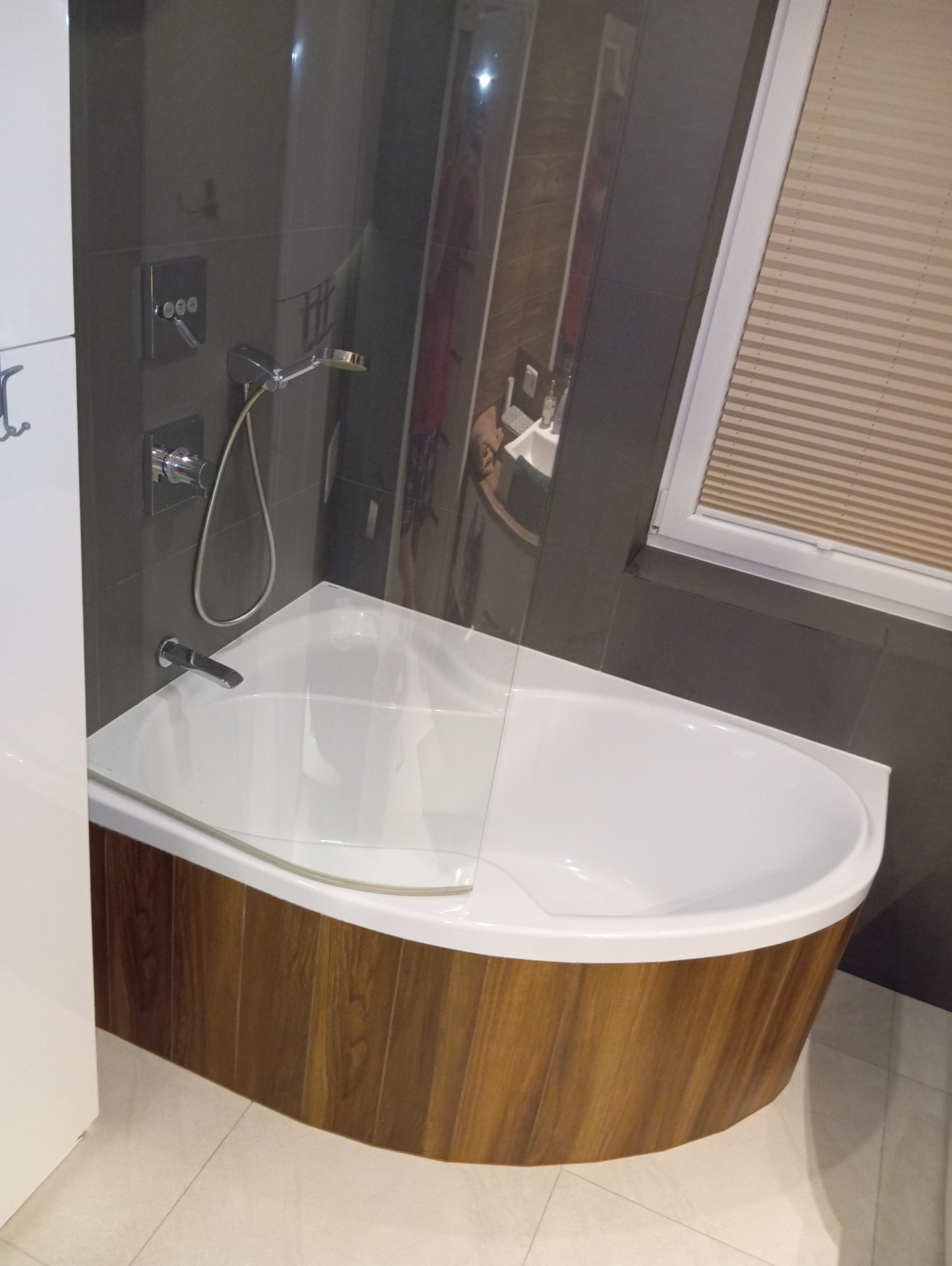 Nowoczesna wanna z drewnopodobnym wykończeniem w łazience w Białymstoku, z szarą glazurą i szklanym parawanem nawannowym, widok na baterię i okno z żaluzjami.