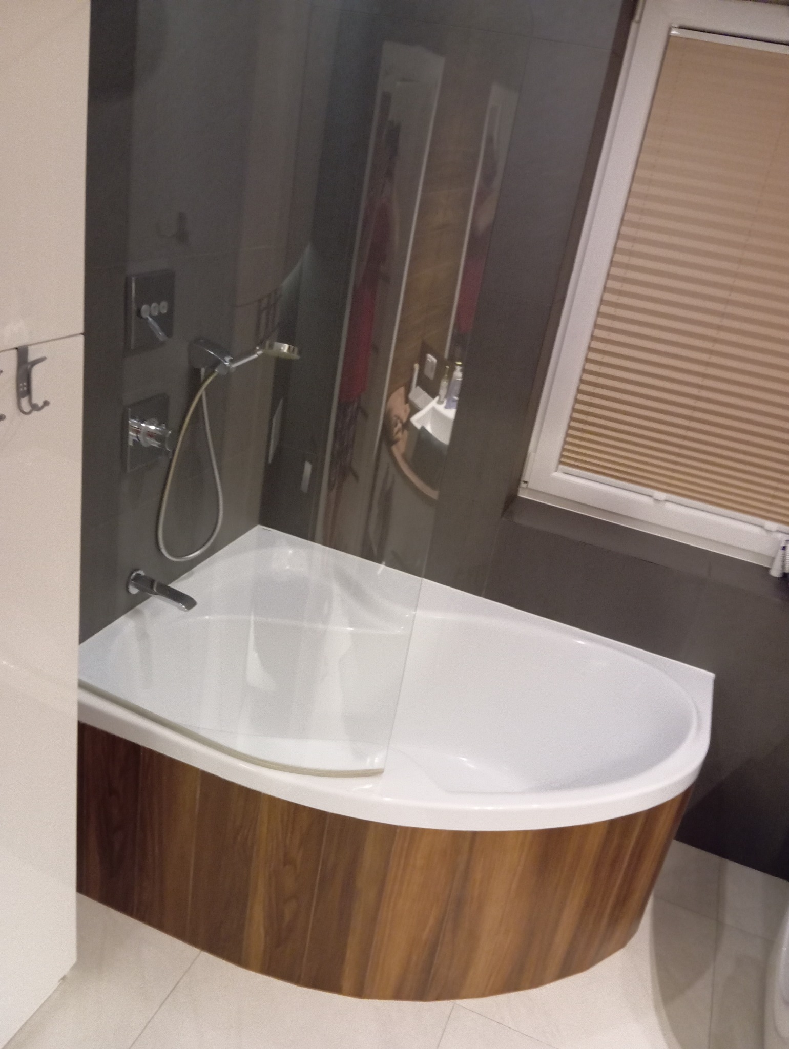 Nowoczesna wanna z obudową imitującą drewno, bateria i deszczownica na ciemnoszarych płytkach, widok na okno z żaluzjami plisowanymi.