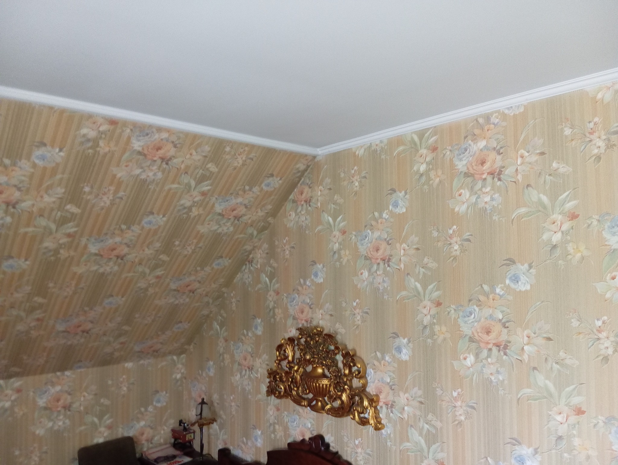 Pokój na poddaszu z tapetą w kwiaty, ukośny sufit i ozdobny, złoty ornament na ścianie.