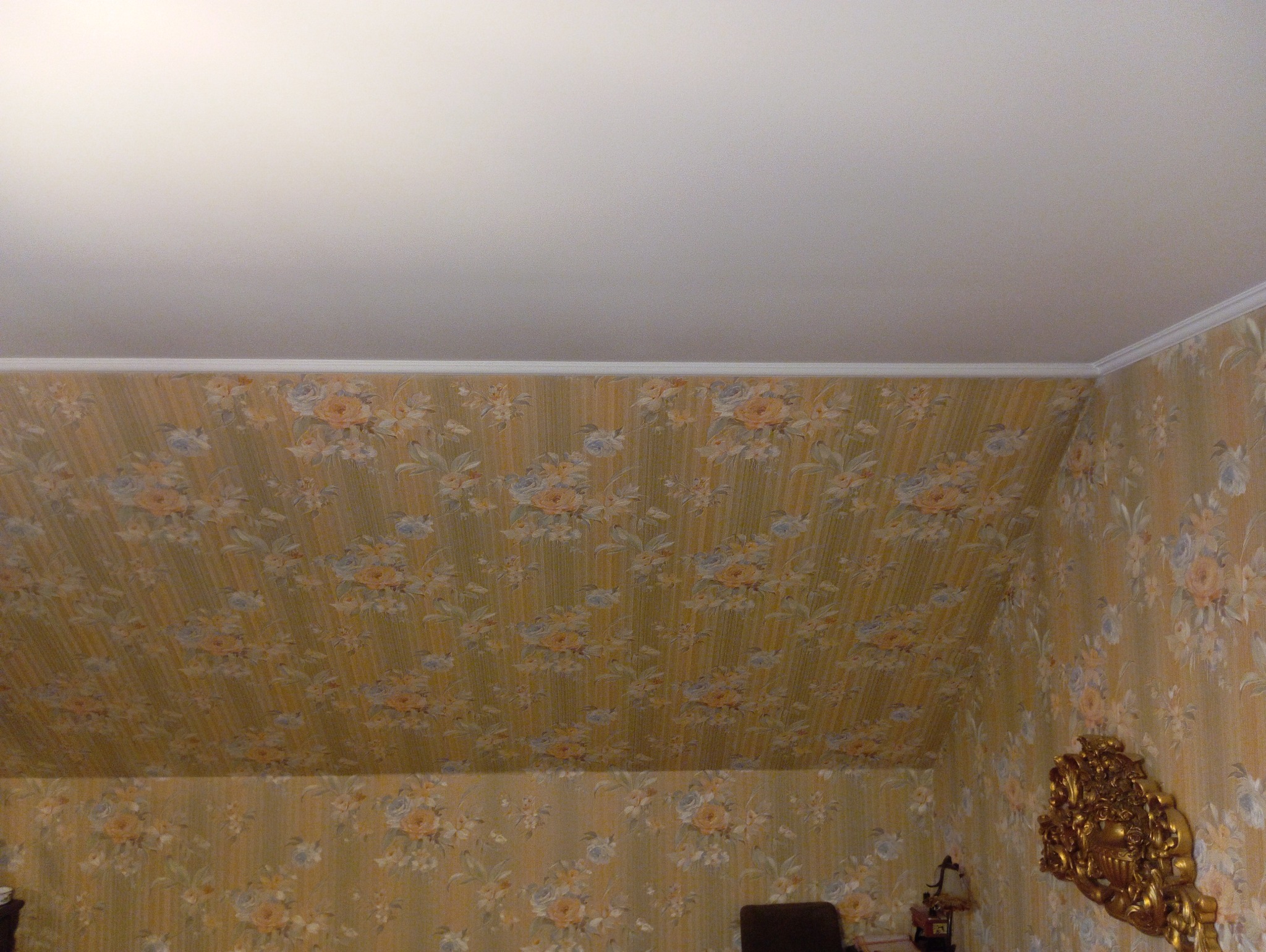 Pokój na poddaszu z żółtą tapetą w kwiaty i białym sufitem, widoczne listwy przypodłogowe i dekoracyjny element na ścianie.