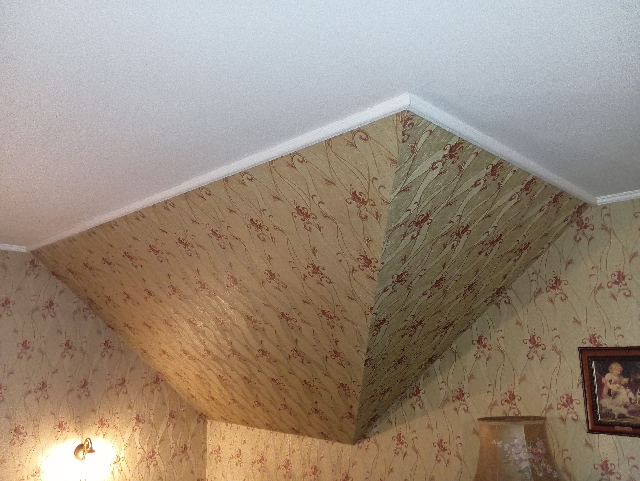 Pokój z beżową tapetą w czerwone kwiaty na ścianach i skośnym suficie, wykończony białym gzymsem.