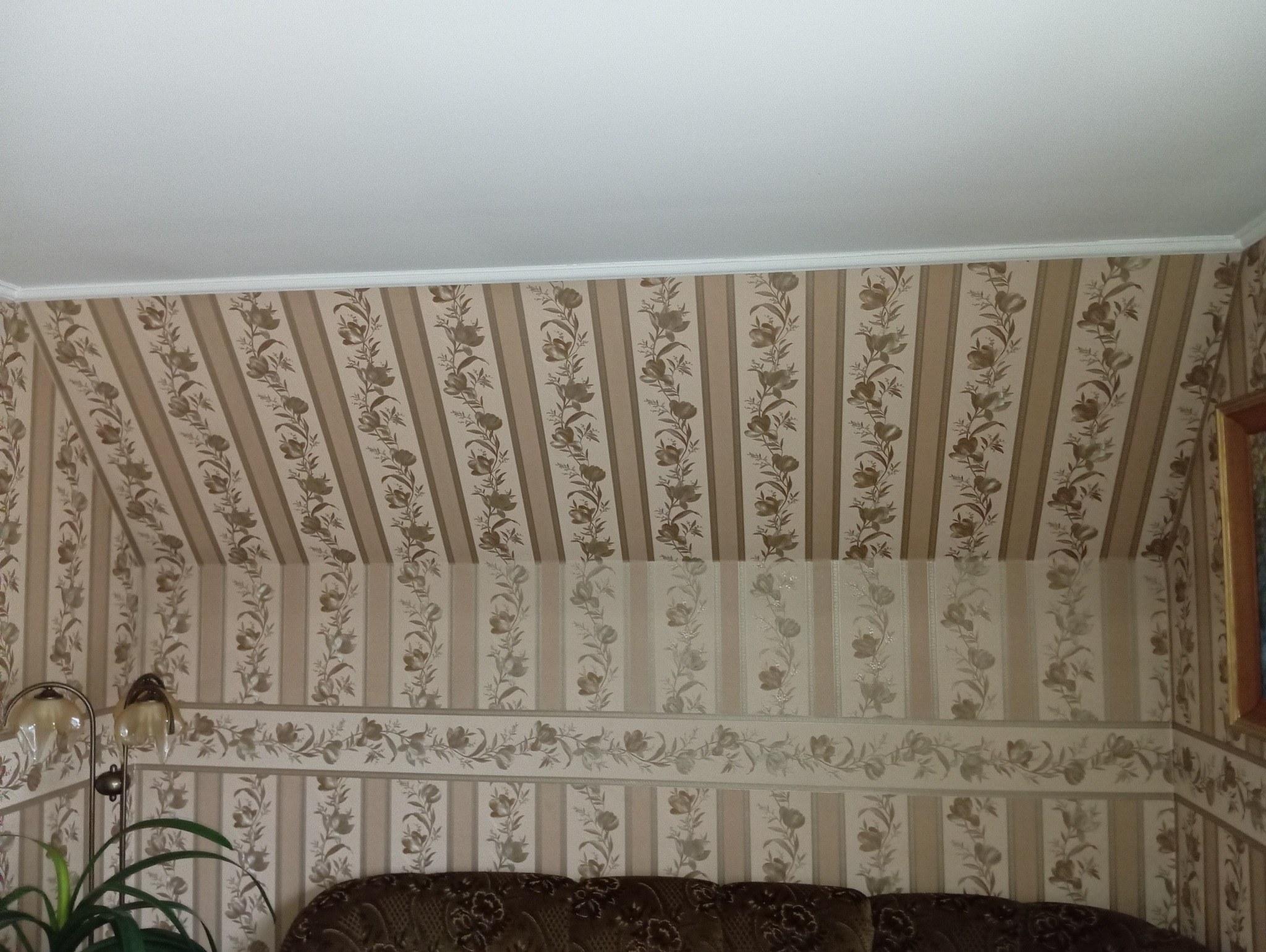 Beżowe tapety w pionowe pasy z motywem kwiatowym zdobią ściany i skośny sufit poddasza, widoczny fragment sofy i lampy.