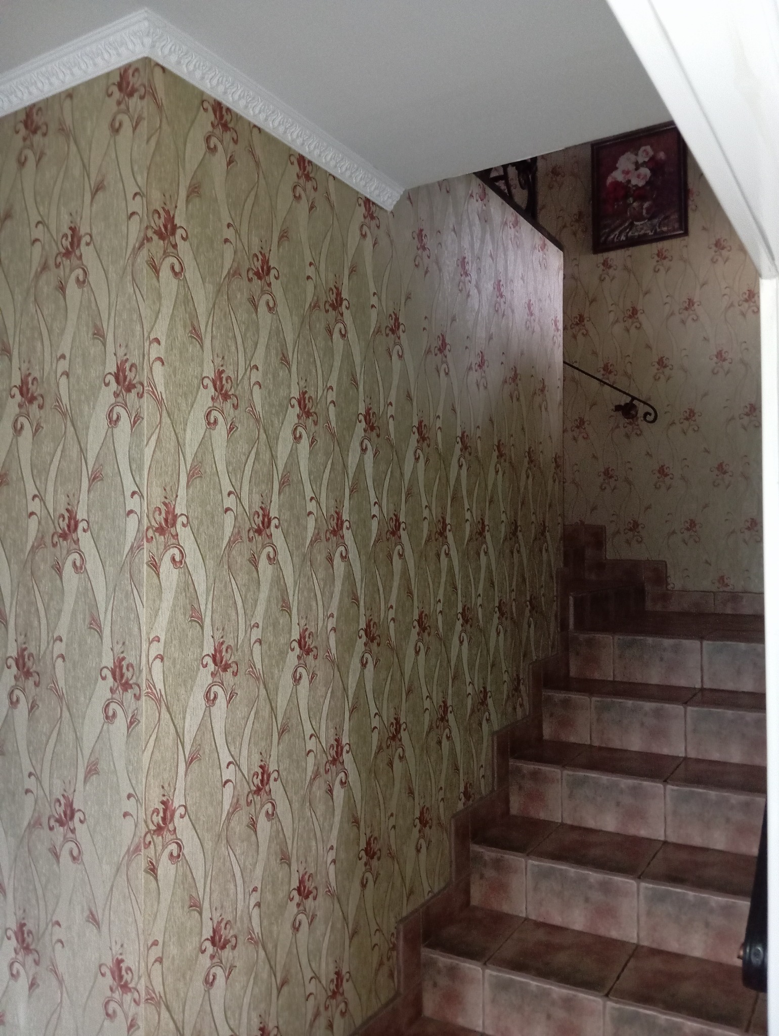 Klatka schodowa z tapetą w kremowo-czerwone kwiaty i brązowymi schodami wyłożonymi kafelkami, ozdobny biały gzyms przy suficie.
