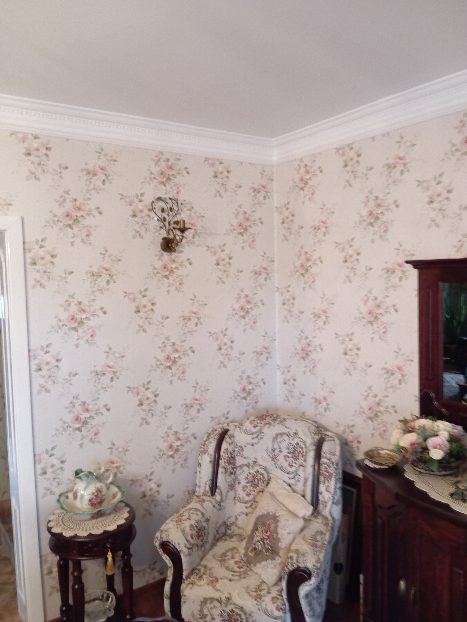 Umeblowane wnętrze pokoju z tapetą w delikatne róże, ozdobnym krzesłem, komodą i dekoracyjnym oświetleniem ściennym.