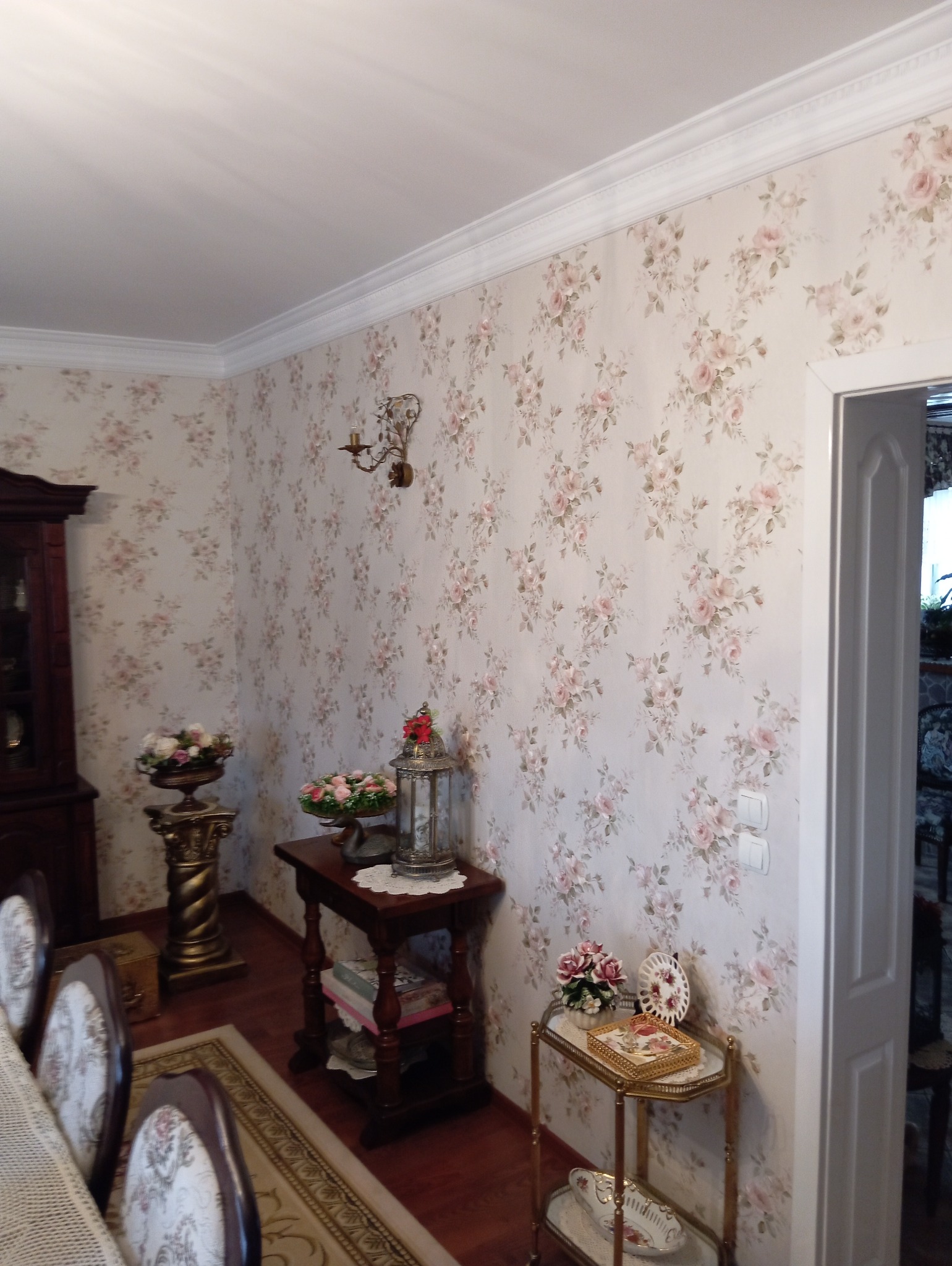Pokój z tapetą w drobne róże, ozdobnymi listwami przysufitowymi, antykami i dekoracjami, widok na fragment jadalni z krzesłami w stylu retro
