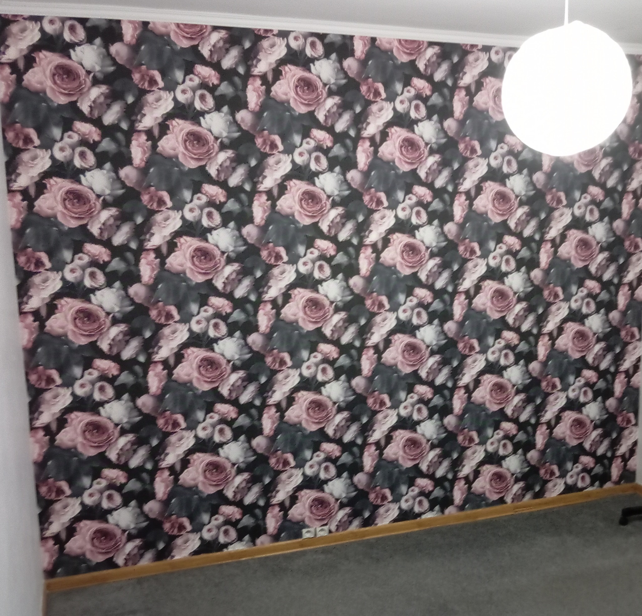 Ściana pokryta tapetą w ciemne, kwieciste wzory z różowymi różami, oświetlona przez kulistą lampę sufitową.
