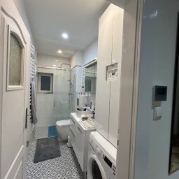 NaVo-Bud - Widok na odnowioną łazienkę z pralką Electrolux i kabiną prysznicową, przez otwarte białe drzwi z dekoracyjnym przeszkleniem. Na podłodze wzorzyste płytki, w tle toaleta i umywalka.