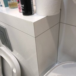 NaVo-Bud - Nowoczesna, biała toaleta podwieszana z zabudową z płytek ceramicznych, na której stoją akcesoria łazienkowe: zapach do pomieszczeń i papier toaletowy.