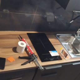 NaVo-Bud - Przygotowanie do montażu płyty indukcyjnej i zlewu w kuchni: widoczny pistolet do silikonu, kartusz silikonu, smartfon i narzędzia na blacie.