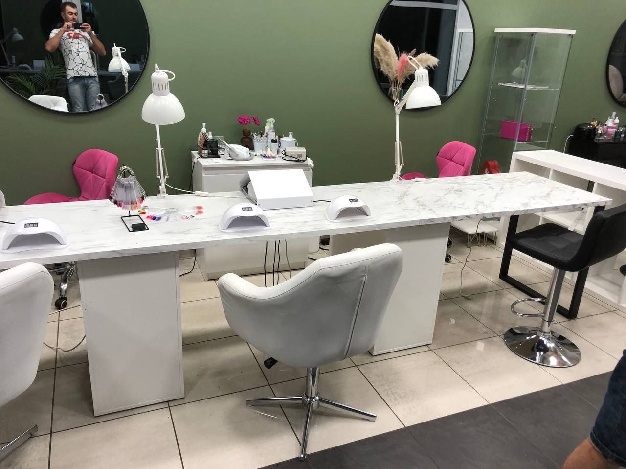 Stanowiska do manicure z lampami UV, marmurowym blatem i różowymi krzesłami na tle oliwkowozielonej ściany z okrągłymi lustrami.