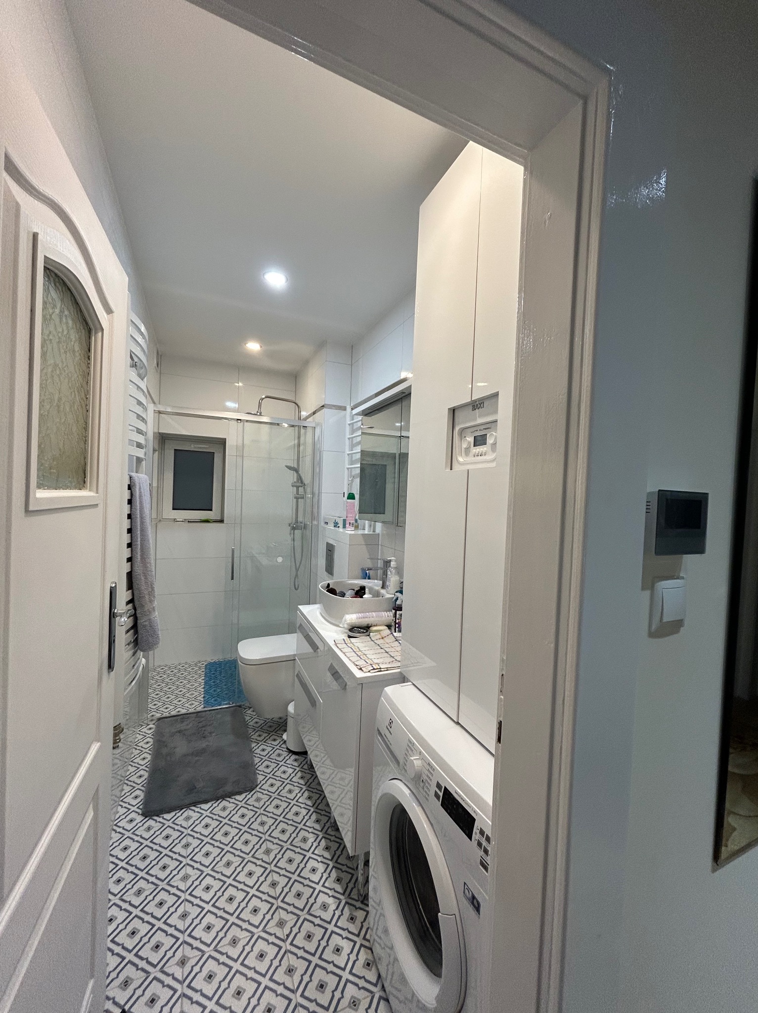 Widok na odnowioną łazienkę z pralką Electrolux i kabiną prysznicową, przez otwarte białe drzwi z dekoracyjnym przeszkleniem. Na podłodze wzorzyste płytki, w tle toaleta i umywalka.