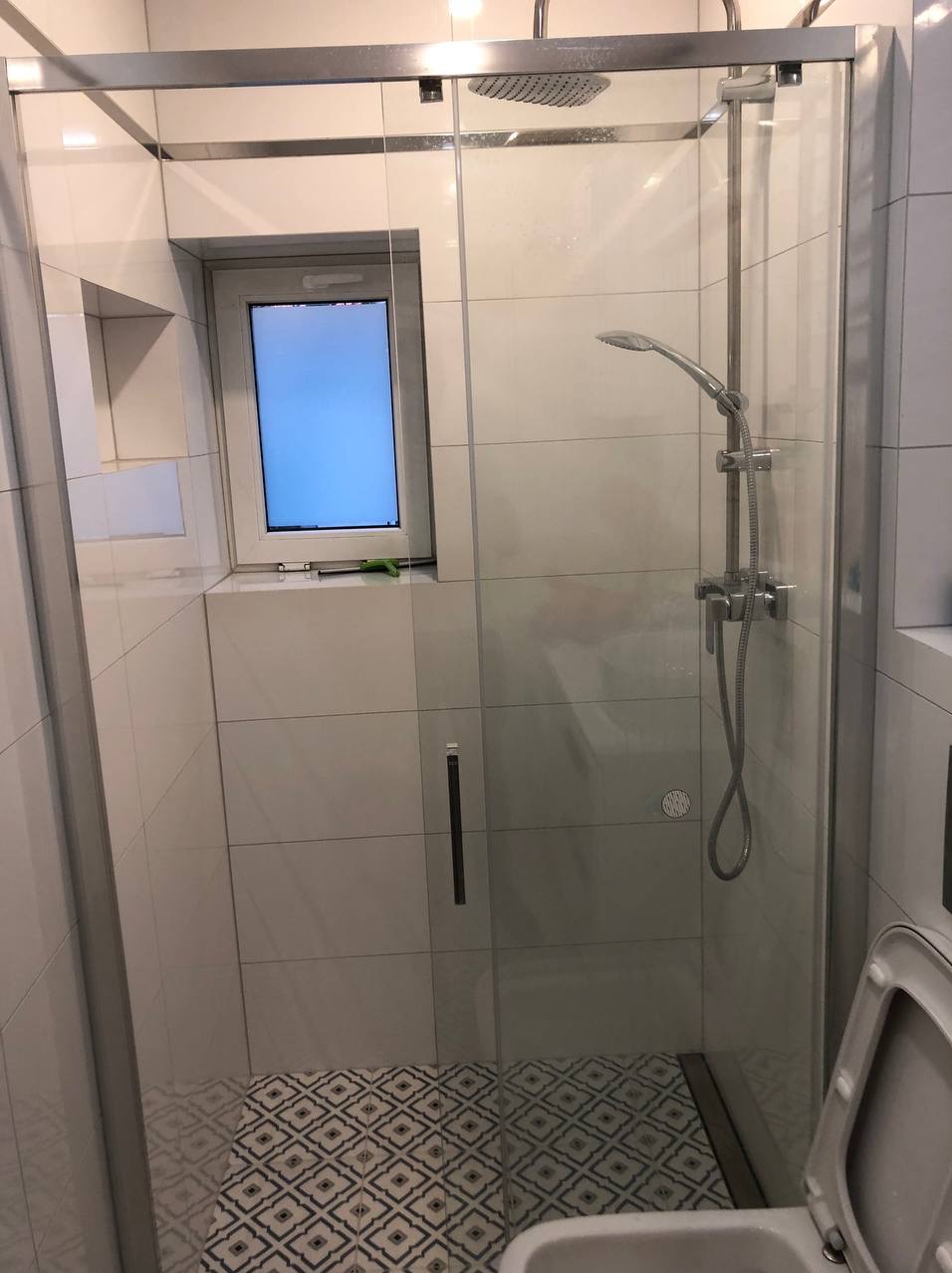 Nowoczesna kabina prysznicowa z deszczownicą i szklanymi drzwiami, jasne płytki ścienne i wzorzysta podłoga, małe okno w kabinie.