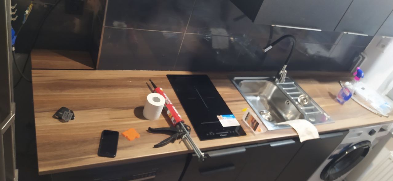 Przygotowanie do montażu płyty indukcyjnej i zlewu w kuchni: widoczny pistolet do silikonu, kartusz silikonu, smartfon i narzędzia na blacie.