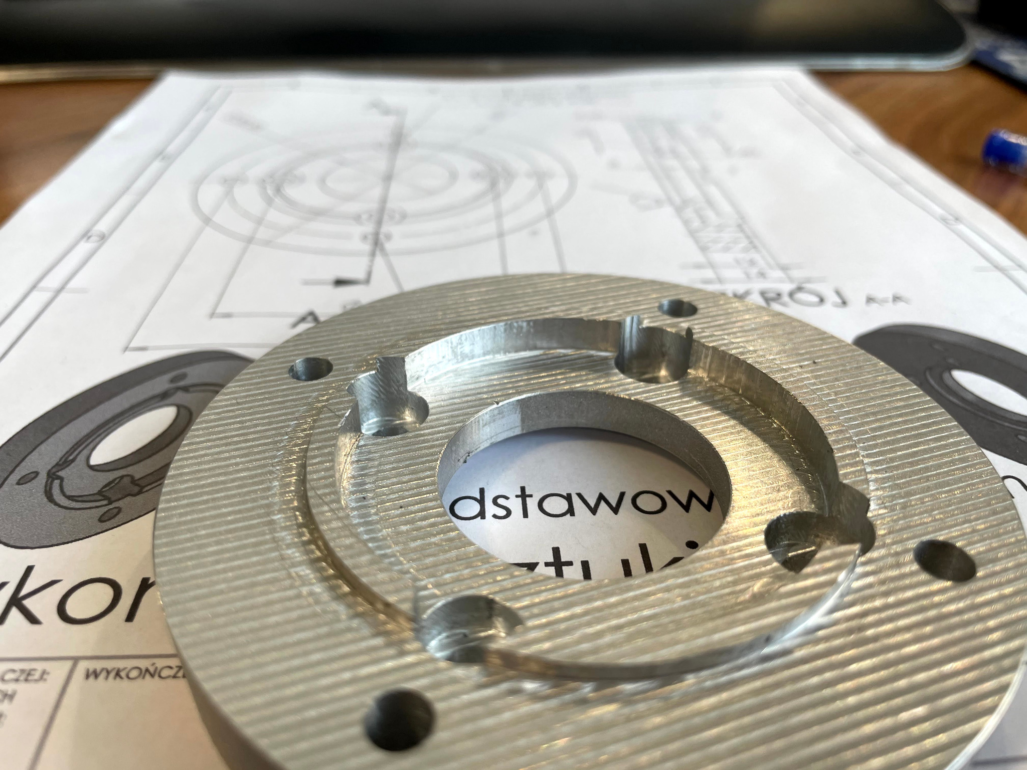 Aluminiowy pierścień z otworami na tle rysunku technicznego, widoczne ślady obróbki skrawaniem.