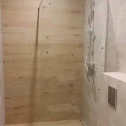 Nowoczesna łazienka z kabiną prysznicową typu walk-in, wykończona płytkami imitującymi drewno, z minimalistyczną toaletą i odpływem liniowym.