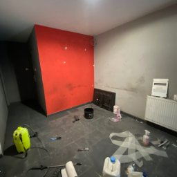 Paweł Pecyna - Pokój w trakcie prac wykończeniowych: czerwona ściana z niedomalowaniami, szara ściana w plamach, na podłodze narzędzia, pojemniki z farbą i folia malarska.