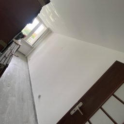 Paweł Pecyna - Świeżo pomalowane na biało wnętrze pokoju w Rogoźniku, widoczna drabina malarska przy oknie i częściowo zamontowane ciemne drzwi.