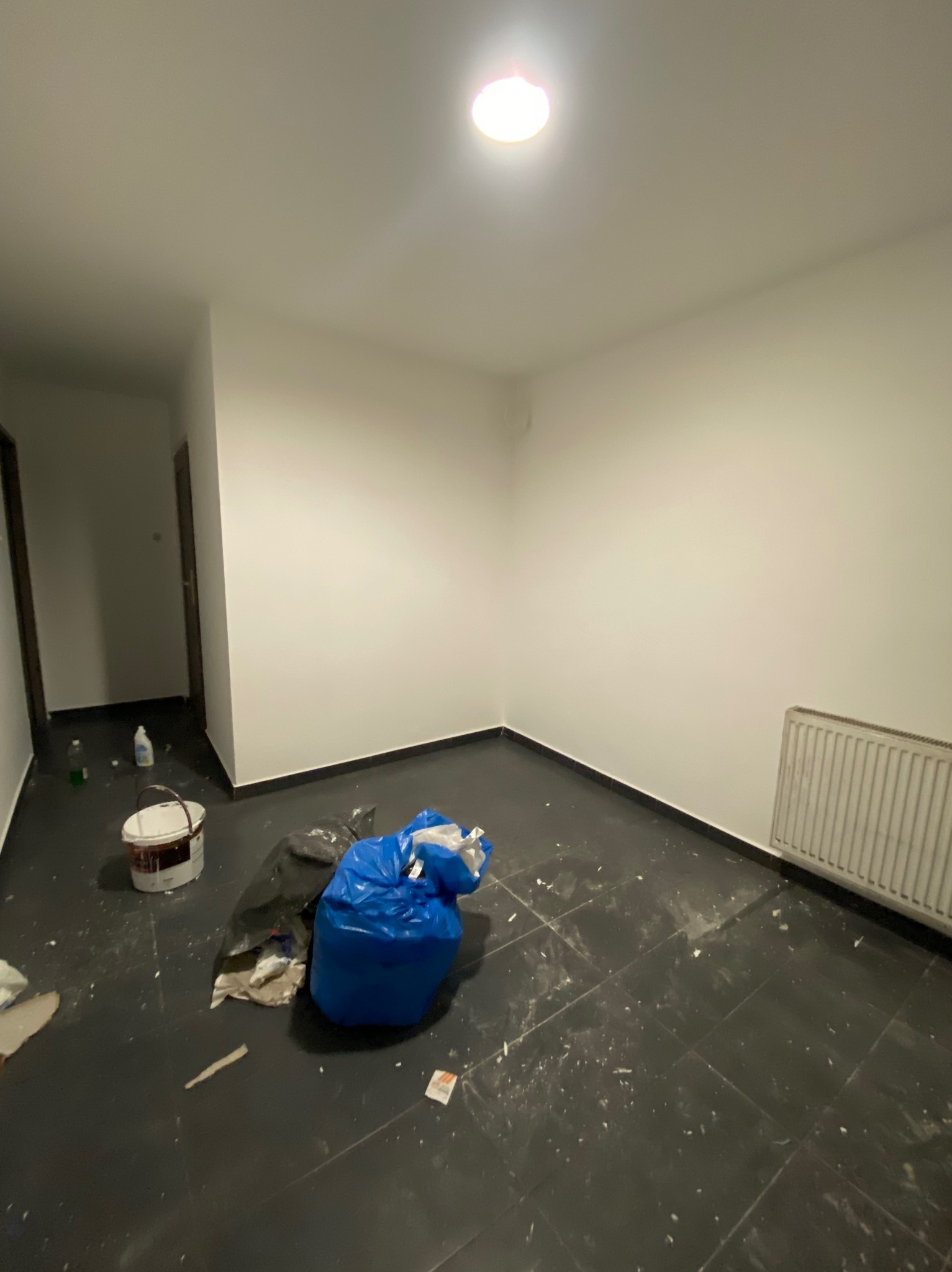Pokój po świeżo zakończonym malowaniu ścian na biało, widoczne ślady farby na czarnej podłodze, wiadro z farbą, worki na śmieci i grzejnik.