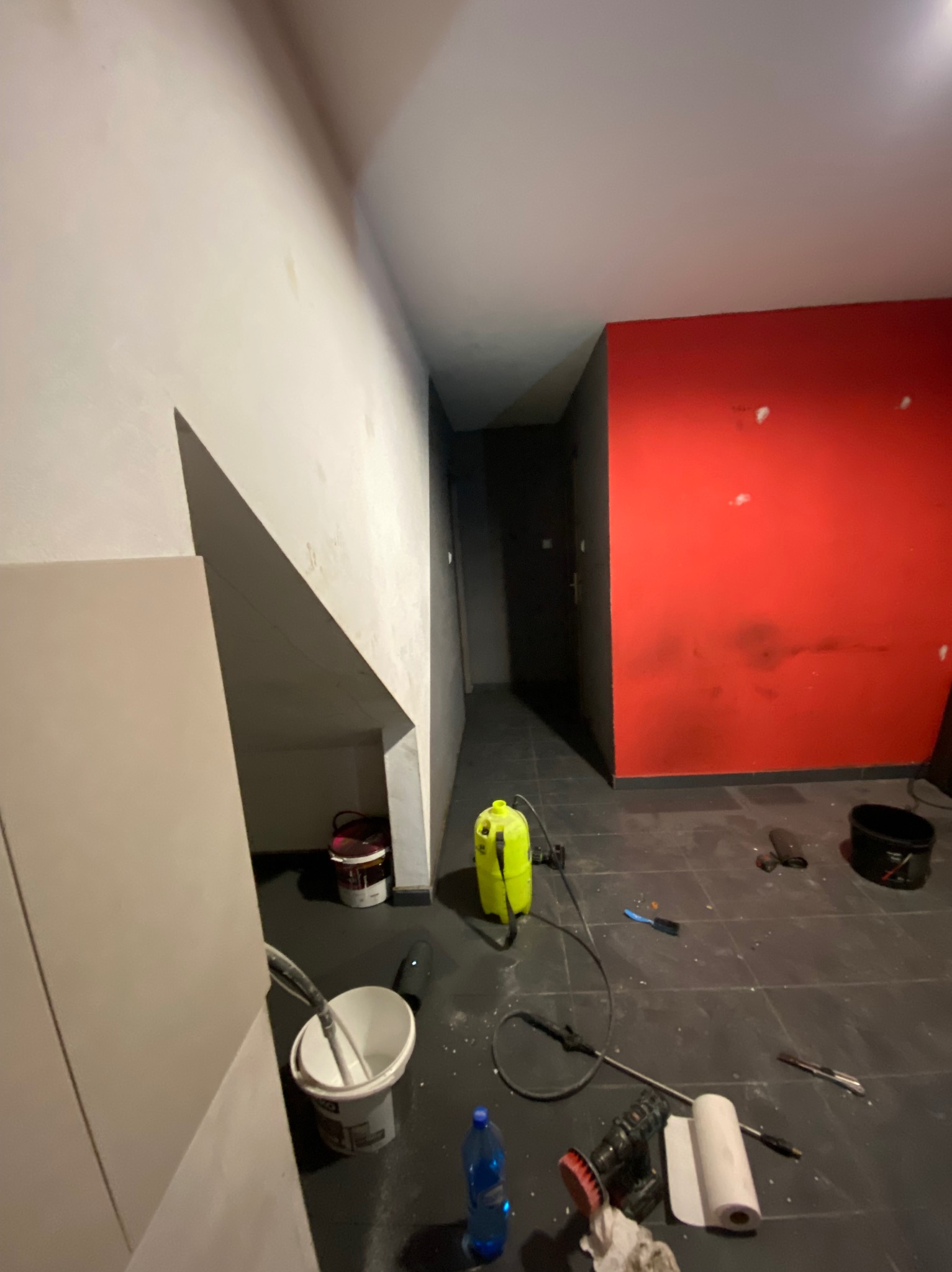 Pokój w trakcie odświeżania: czerwona ściana z widocznymi śladami po szpachlowaniu, biała ściana częściowo pokryta szpachlą, na podłodze narzędzia malarskie, wiadro z mieszadłem i butelka wody.