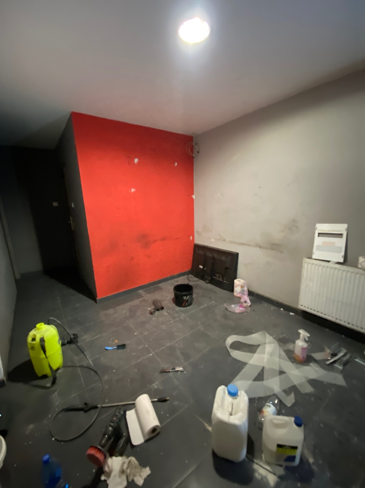 Pokój w trakcie prac wykończeniowych: czerwona ściana z niedomalowaniami, szara ściana w plamach, na podłodze narzędzia, pojemniki z farbą i folia malarska.