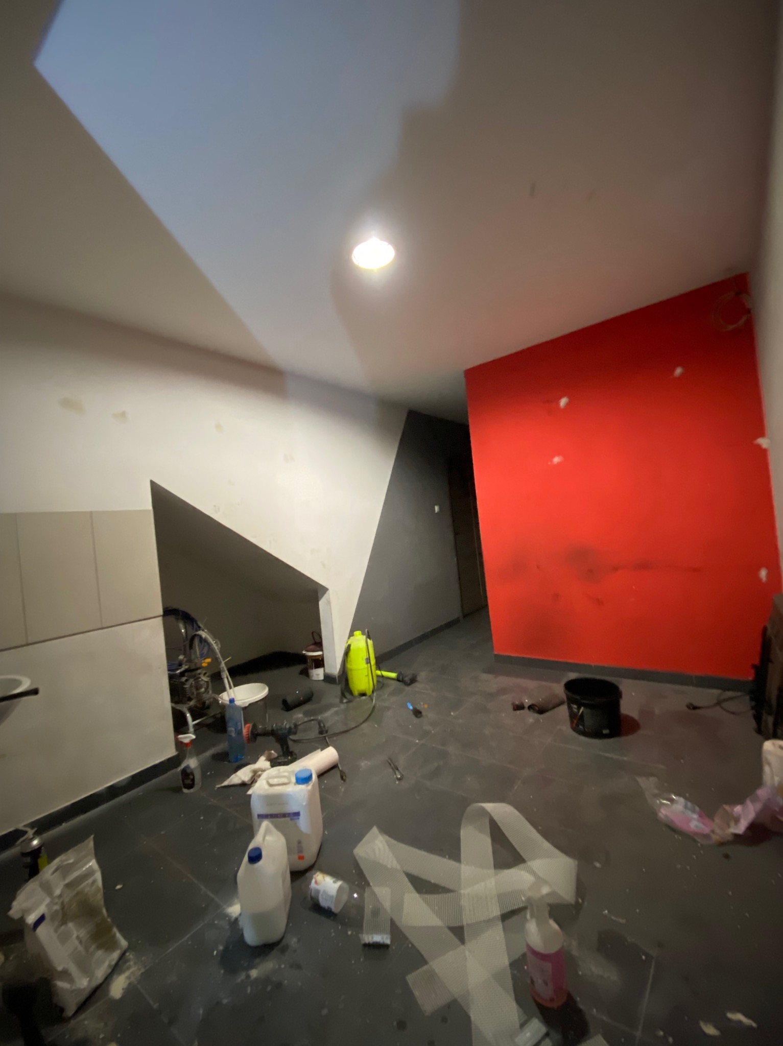 Wnętrze w trakcie odświeżania: ściana pomalowana na intensywny czerwony kolor kontrastuje z szarą ścianą, widoczne narzędzia i materiały budowlane, podłoga zabezpieczona folią malarską...
