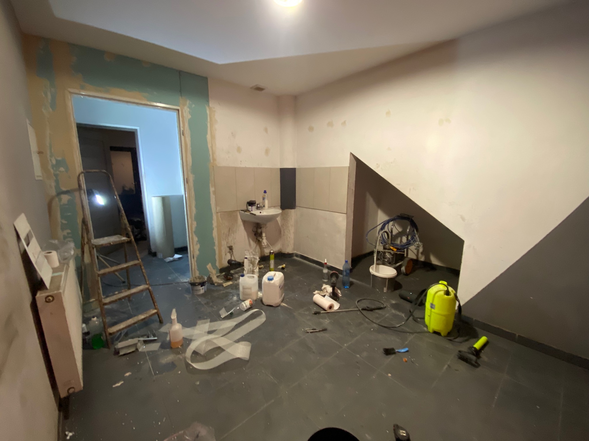 Pokój w trakcie remontu: ściany częściowo otynkowane, drabina, narzędzia malarskie, agregat malarski, resztki materiałów budowlanych na podłodze z płytek.